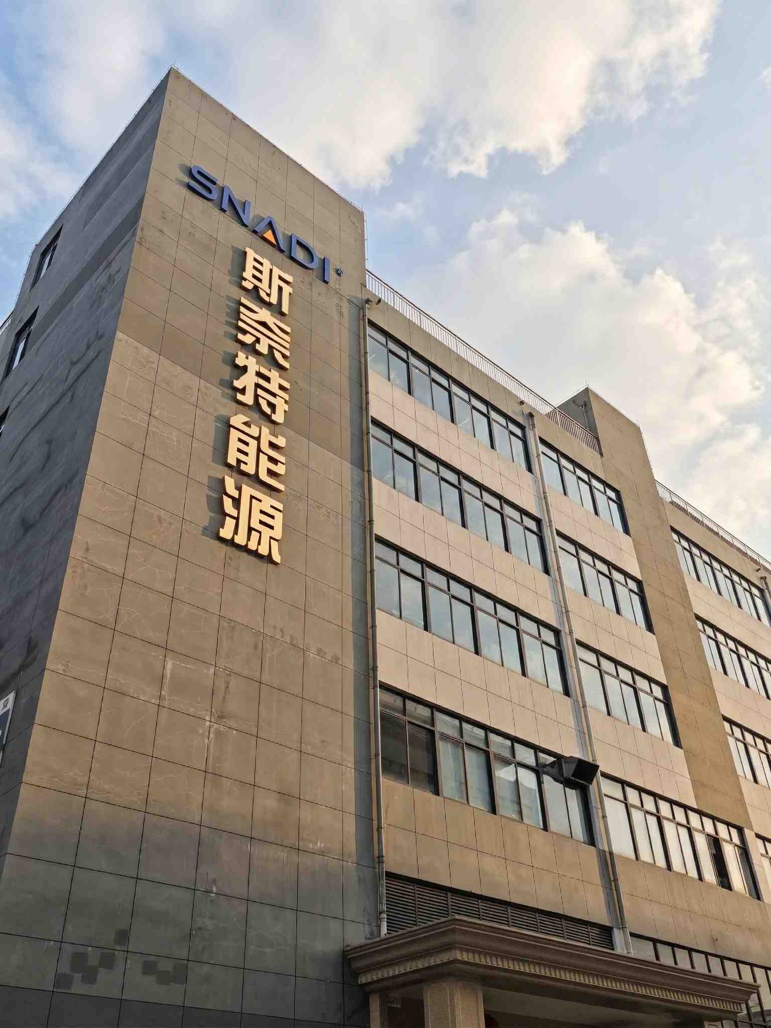 A modern multi-story SNADI renewable energy solutions company building in gray, with the SNADI logo at the top and the golden Chinese characters 斯奈特能源 below, against a partly cloudy sky.